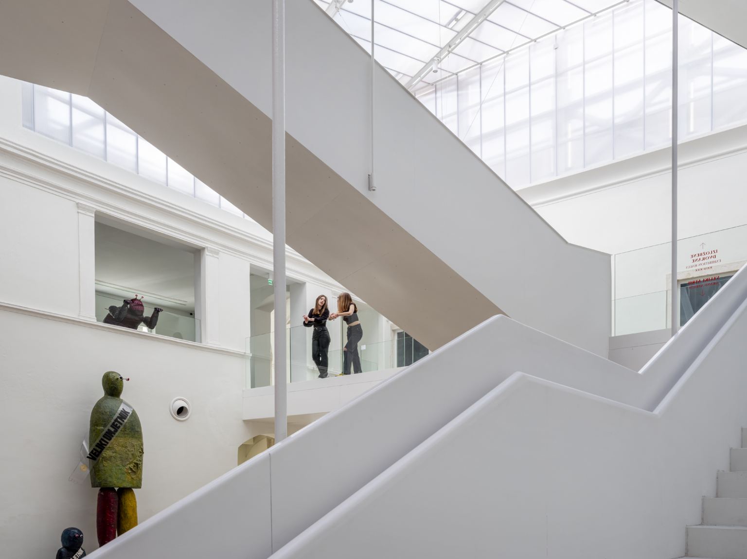 The white daylit atrium of II Palaces, a broad staircase rising beneath a glazed roof past sculptures and the entrance to the exhibition halls