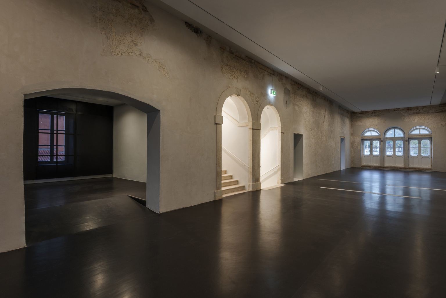 A restored hall in II Palaces, bare plaster walls carrying fragments of historic fresco above a dark polished floor, with arched doorways and tall arched windows along one side