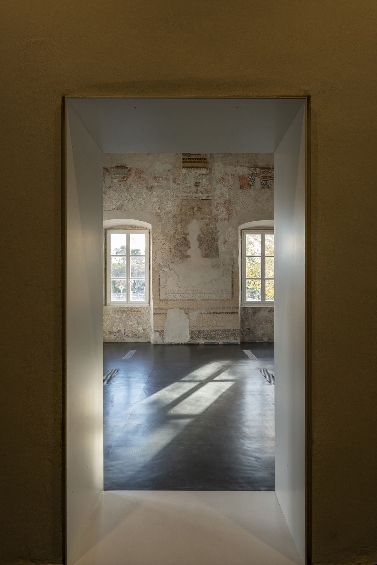 Looking through a doorway into a frescoed room at II Palaces, two arched windows and worn painted plaster catching the afternoon light